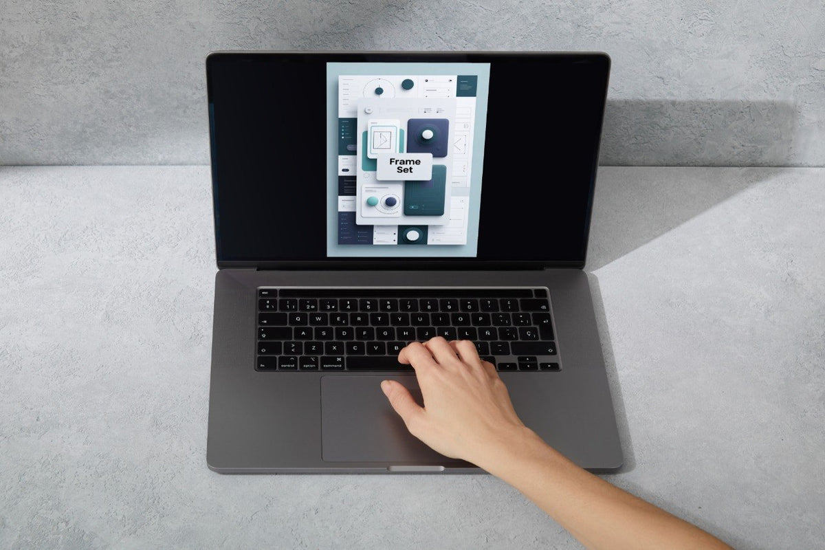 A gray laptop on a light gray surface with a hand resting on the trackpad. The laptop screen displays a digital template preview labeled 'Framer Set'.