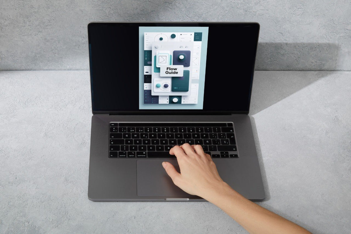 A gray laptop sits on a light gray surface with a hand resting on the trackpad. The screen displays a software interface or digital guide.